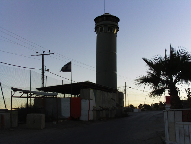 photo où on voit une tour militaire israélienne, entourée de blocs de bétons, un palmier sur le côté droit de la route-entrée