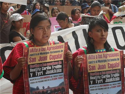 Deux jeunes femmes du village, vêtues de leur costume traditionnelle rouge, tiennent des affiches-posters au sujet des gens assassinés. Il y a une manifestation en appui derrière elles.