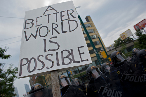 [image: une pancarte dit: Another World Is Possible, mais on voit l'antiémeute en fonds derrière]